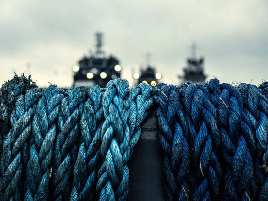 Close-up of textured blue ropes coiled on a bollard with out-of-focus ships in the background under an overcast sky.
