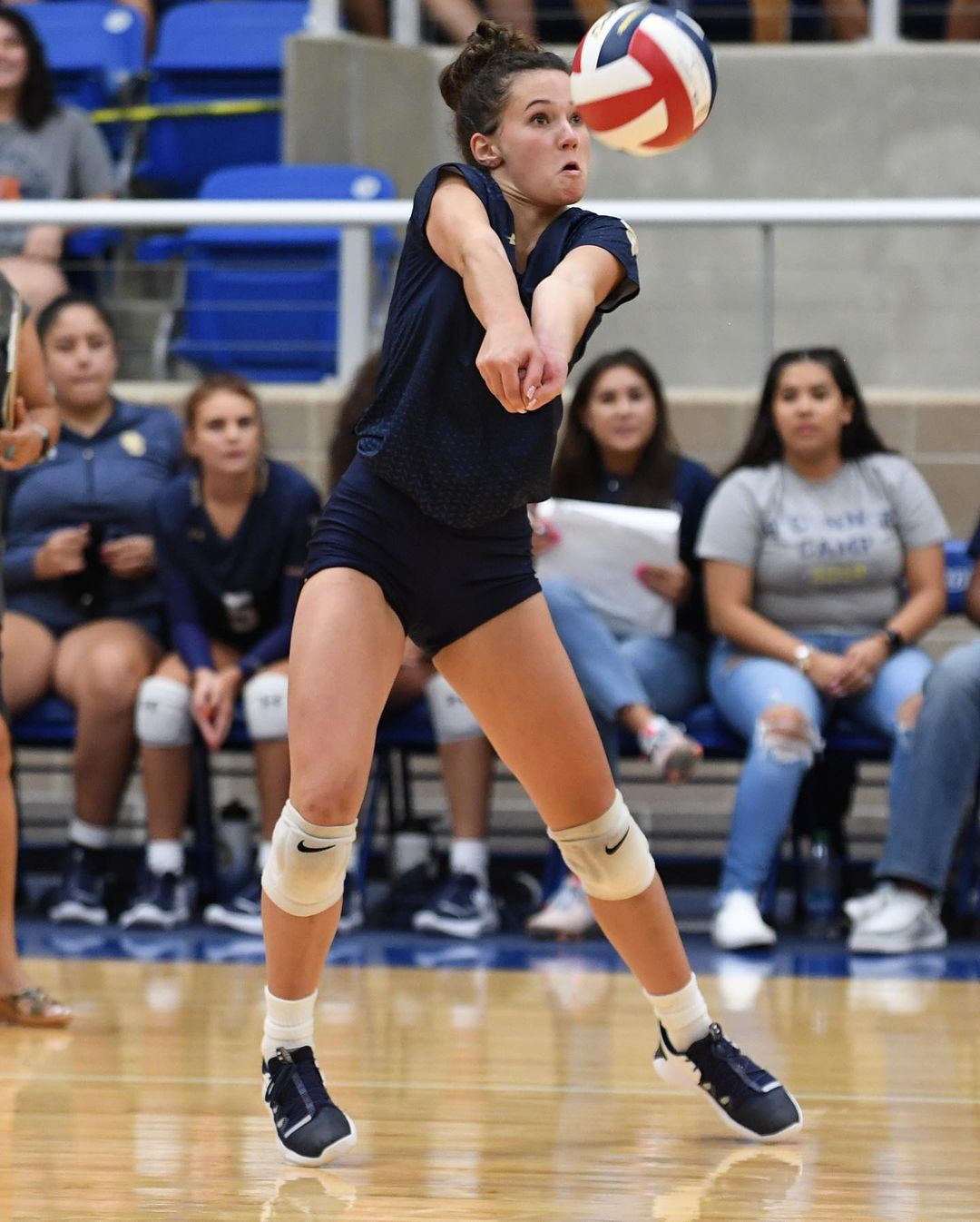 A young woman in sportswear hits a volleyball during an indoor game, focusing with determination in a brightly lit gymnasium setting.