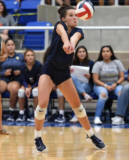 A young woman in sportswear hits a volleyball during an indoor game, focusing with determination in a brightly lit gymnasium setting.