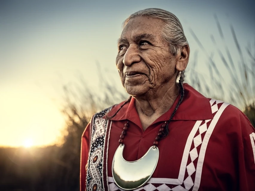Portrait of an elderly Indigenous man wearing traditional clothing, including a silver neck piece and beaded necklace, against an outdoor background.