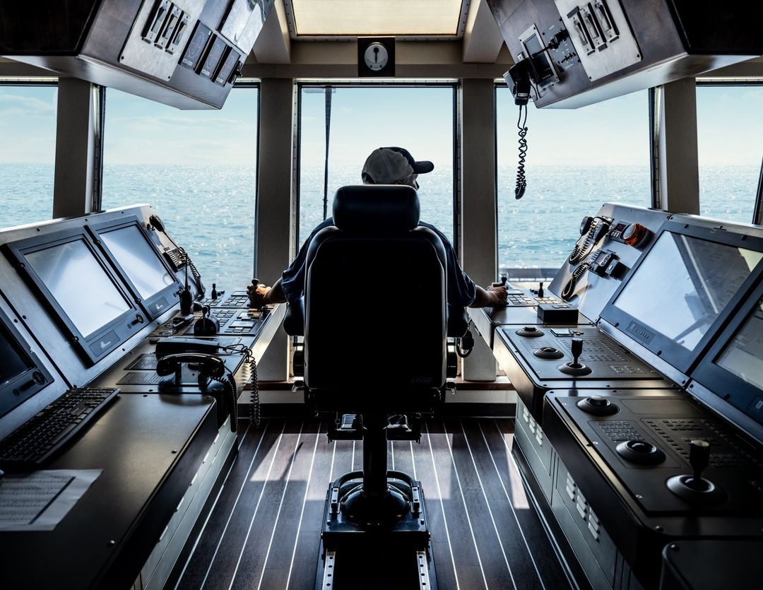 Man navigates vessel from ship bridge featuring control panels, view of the sea through windows, and dark wood flooring in the interior space.