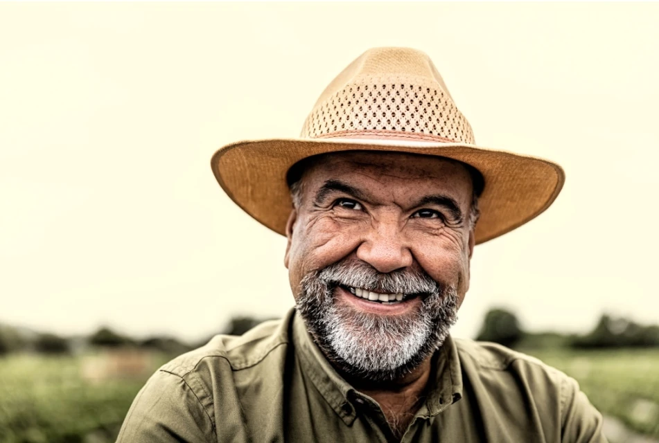 Close-up of a smiling elderly man with a grey beard wearing a straw hat and green shirt, with a blurred background of green foliage.