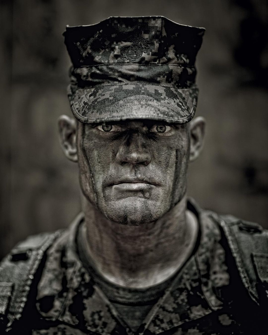 A close-up portrait of an adult male in military uniform and cap, face partially covered in dark camouflage paint, serious expression.