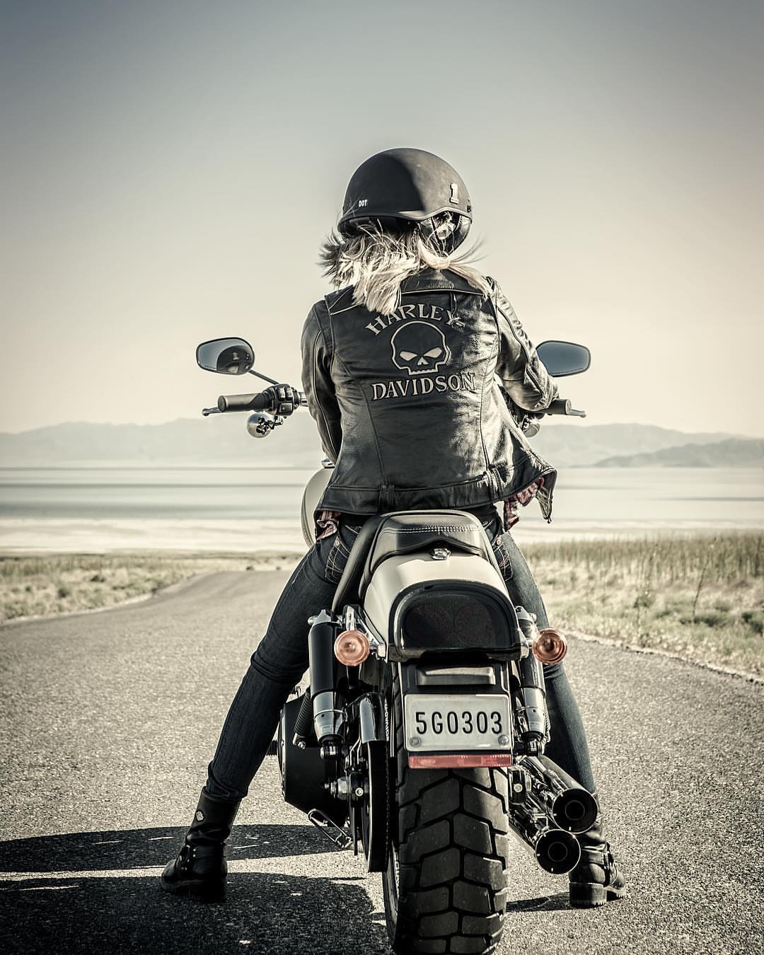 Back view of a woman riding a Harley-Davidson motorcycle on a desert road wearing a helmet, leather jacket, and boots, with a mountain backdrop.