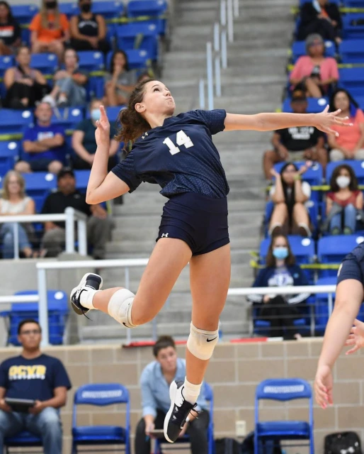 A female volleyball player in mid-air, wearing a navy jersey with number 14, preparing to spike the ball during an indoor match.