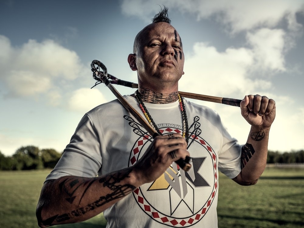 Adult man with mohawk and lacrosse sticks posing outdoors against sky and field background wearing t-shirt with graphic design.