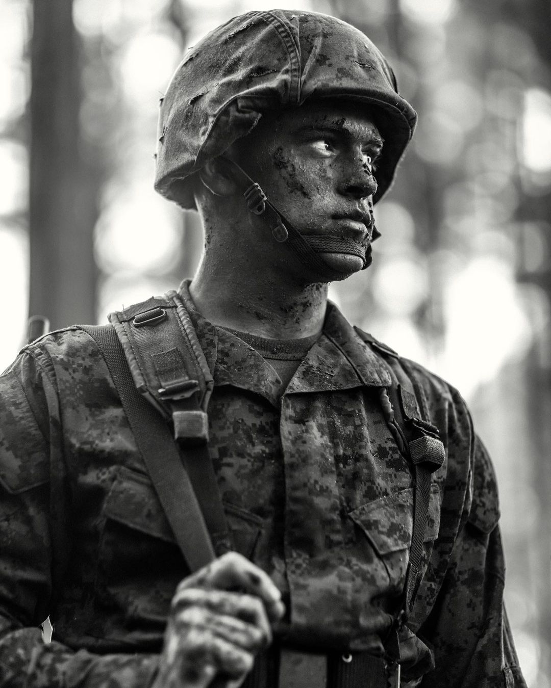 Monochrome portrait of a serious soldier in camouflage uniform and helmet with mud on his face, outdoors in a wooded area.