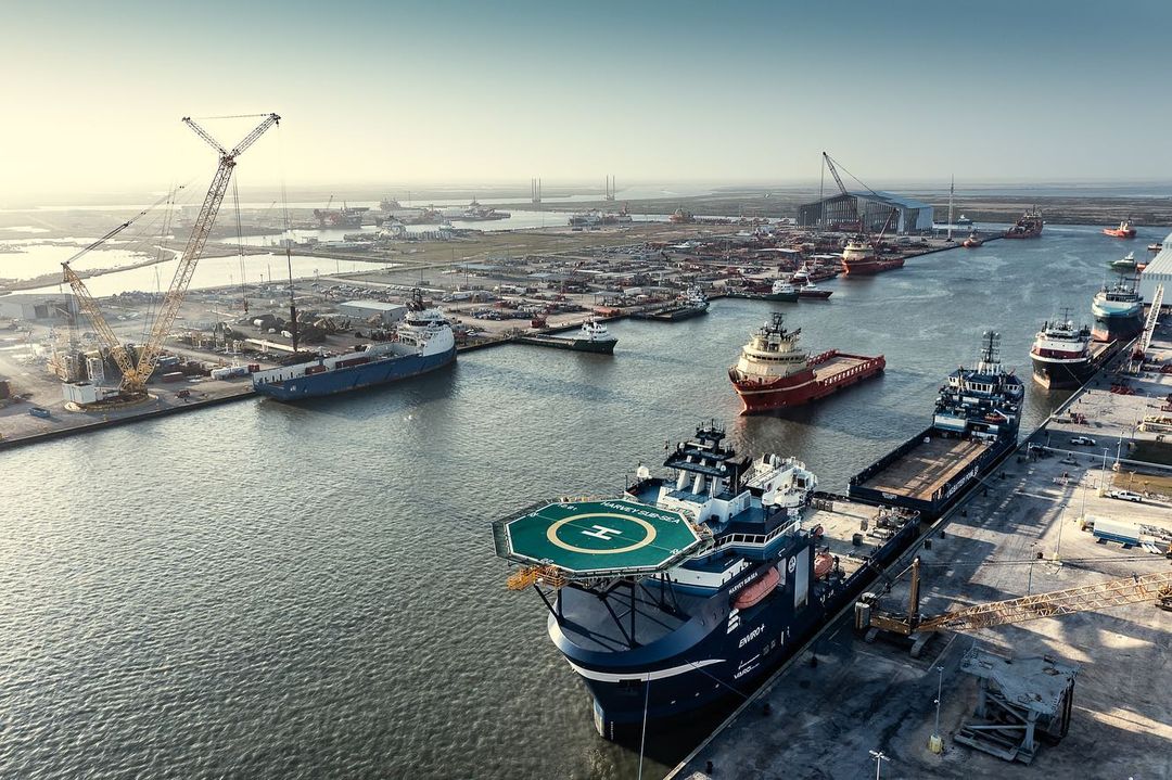 Aerial view of several offshore supply vessels docked at a large industrial port with cranes, buildings, and other ships in the background.