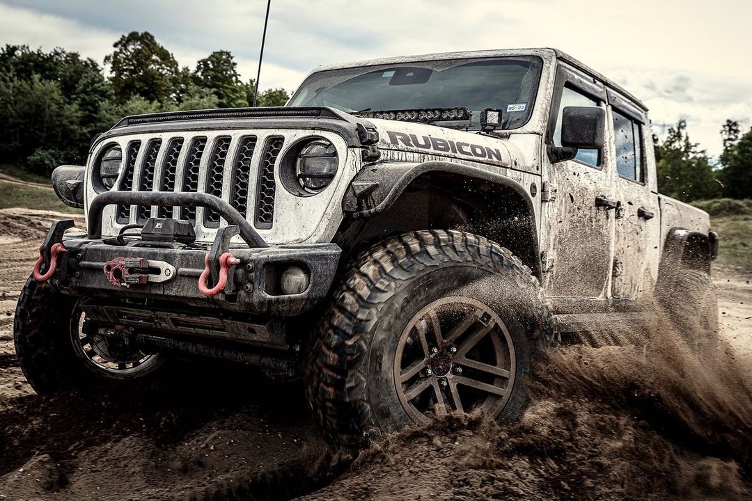 White Jeep Gladiator Rubicon navigating a muddy off-road course with trees visible in the background under a cloudy sky.