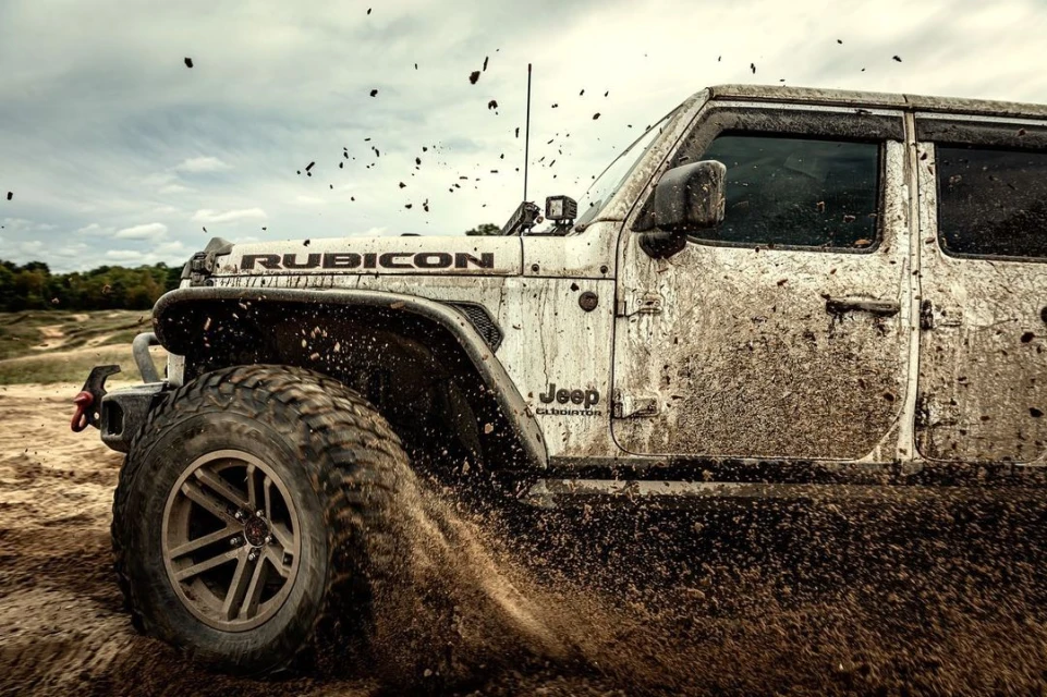 White Jeep Gladiator Rubicon is covered in mud in an off-road action shot on a cloudy day with trails visible in the background.