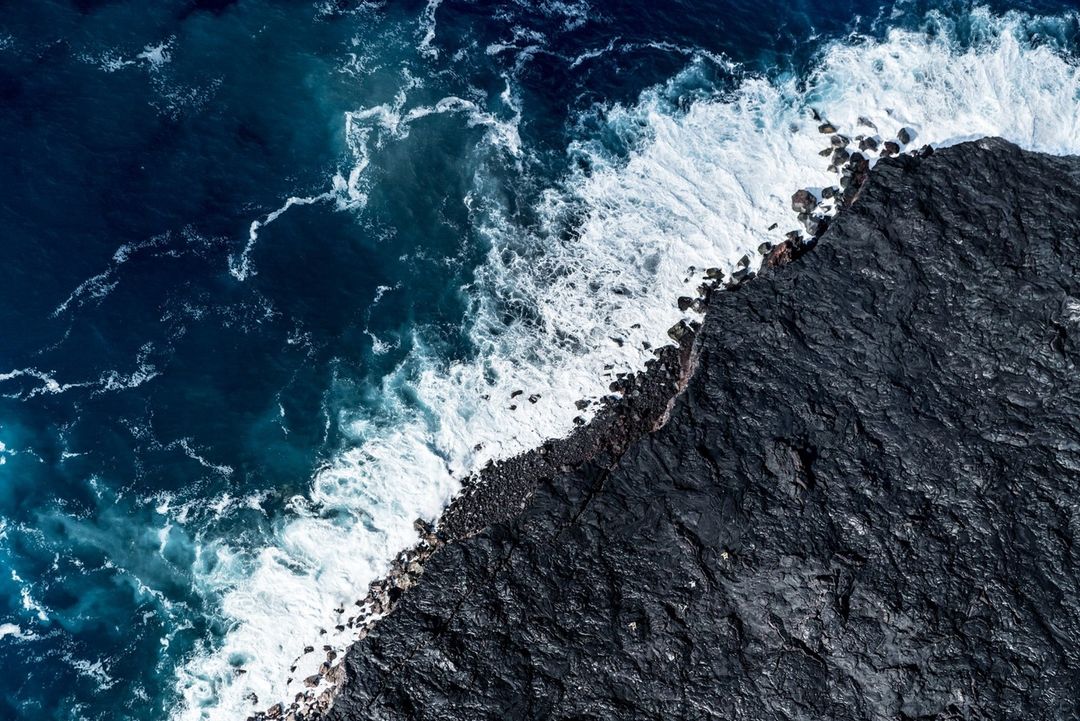Aerial view of the ocean waves crashing against a rocky shoreline. Dark blue water with white foam contrasts with the dark rocks.
