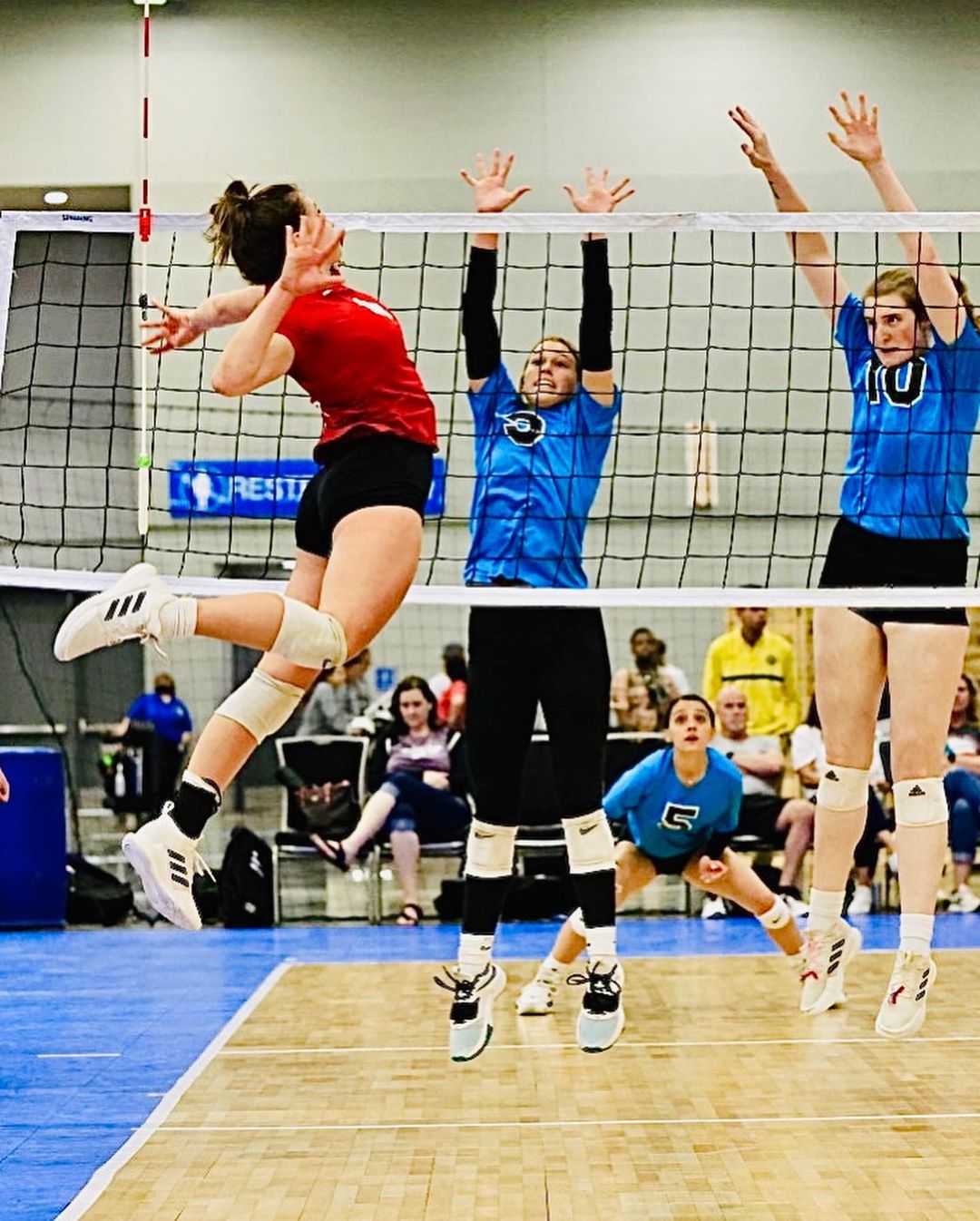 Indoor volleyball action shot featuring a female player in red mid-spike, defenders jumping at the net, and spectators in the background.