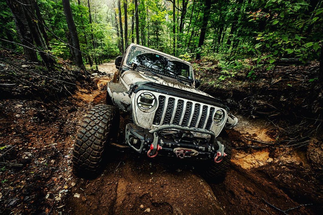 Off-road vehicle navigates a muddy trail through a lush green forest. The vehicle is covered in mud, showcasing its off-roading capabilities.