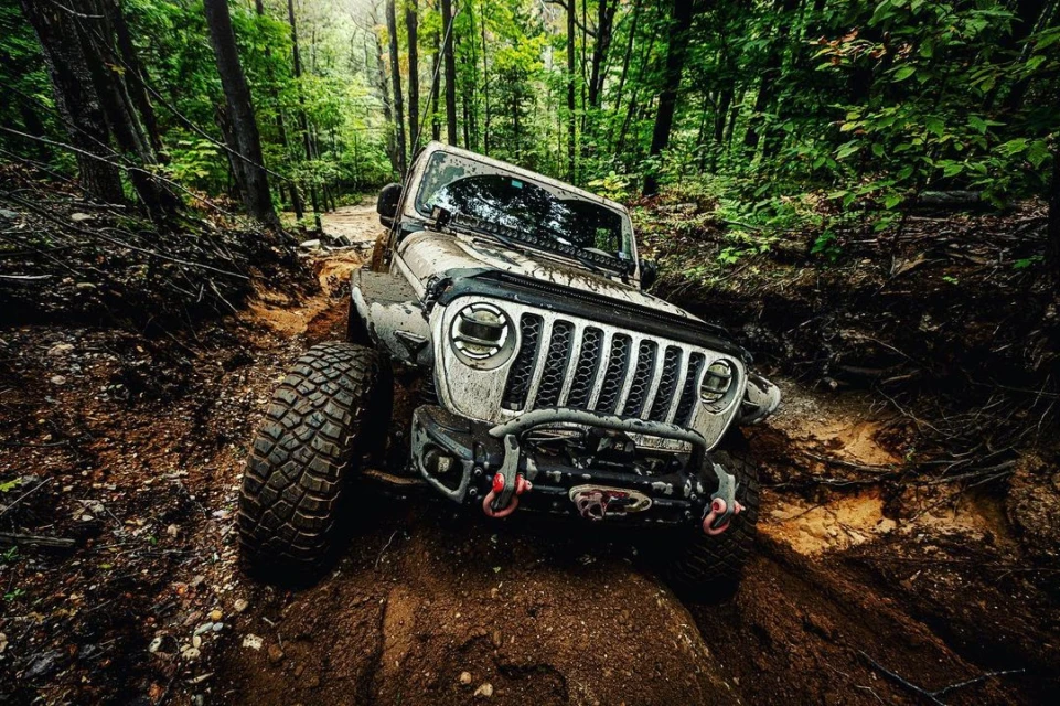 Off-road vehicle navigates a muddy trail through a lush green forest. The vehicle is covered in mud, showcasing its off-roading capabilities.