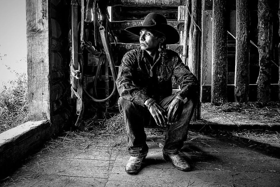 Black-and-white portrait of a man in a cowboy hat sitting in a barn, looking left with his hands resting on his lap.