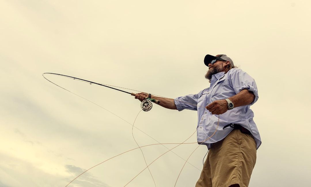 Adult male fly fishing with rod and reel wearing a cap and sunglasses against a cloudy sky in an outdoor environment.