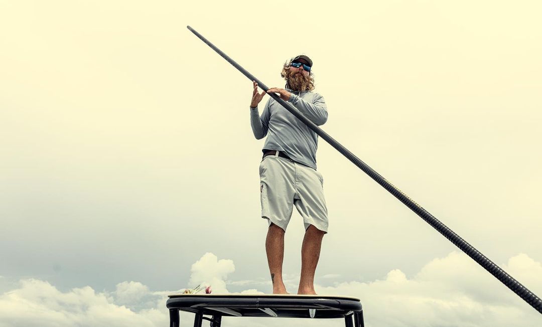 Bearded man stands barefoot on a platform, holding a long black pole against a cloudy sky. He wears sunglasses, a cap, shorts and a long sleeve shirt.