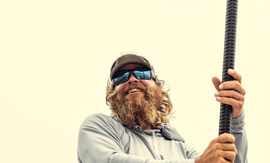 Low-angle view of a smiling adult male with a beard and sunglasses, holding a black pole against a bright, neutral background.