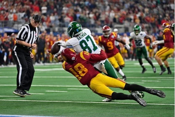 American football action shot: two players in a tackle on a green field during a game, captured in bright daylight.