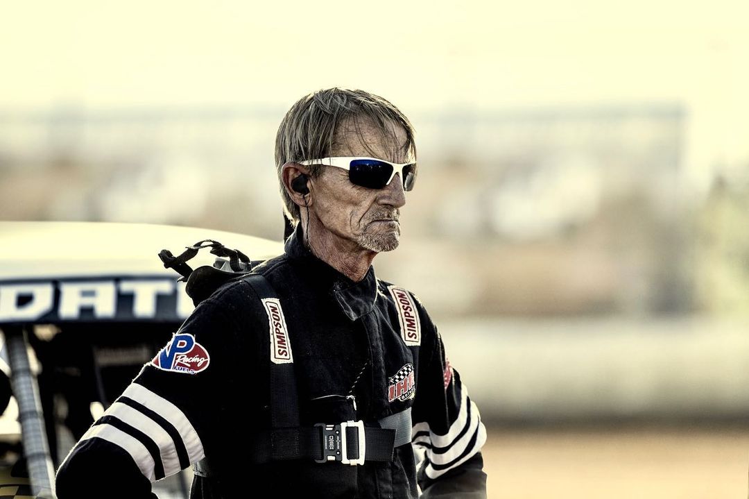 Older adult racer in black racing suit with logos, sunglasses and headset standing next to race car outdoors on a dirt racing track.