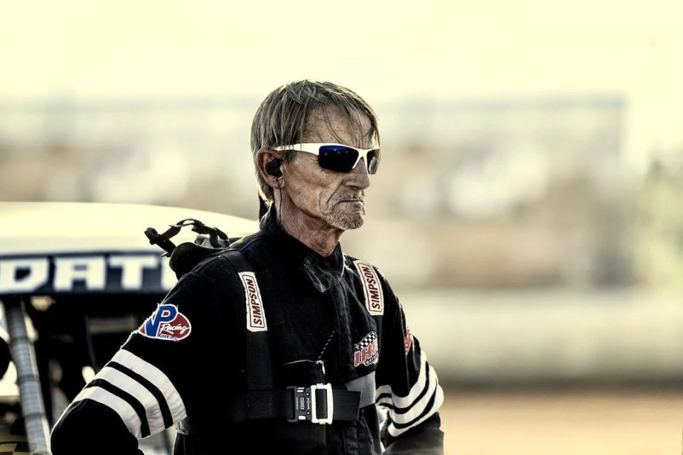 Older adult racer in black racing suit with logos, sunglasses and headset standing next to race car outdoors on a dirt racing track.
