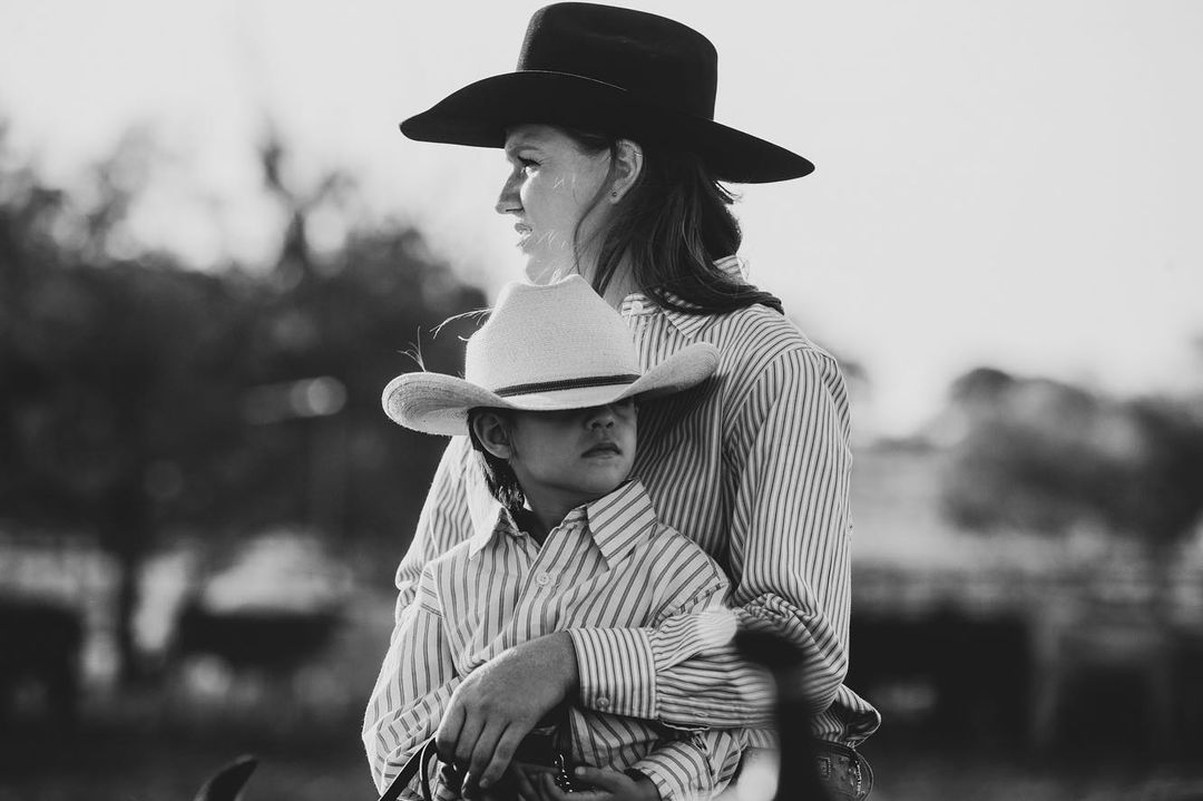 Black and white portrait of a mother and child embracing, wearing cowboy hats and striped shirts in an outdoor setting.