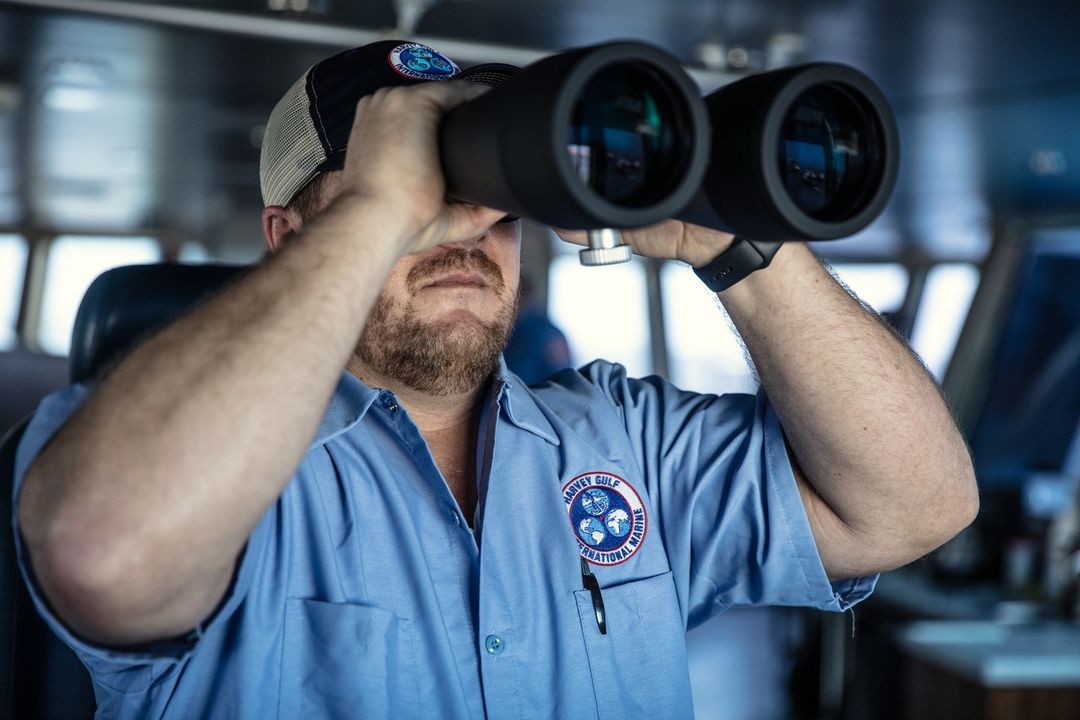 Man in blue work shirt and cap looking through black binoculars in a boat cockpit, concentrating on maritime navigation.