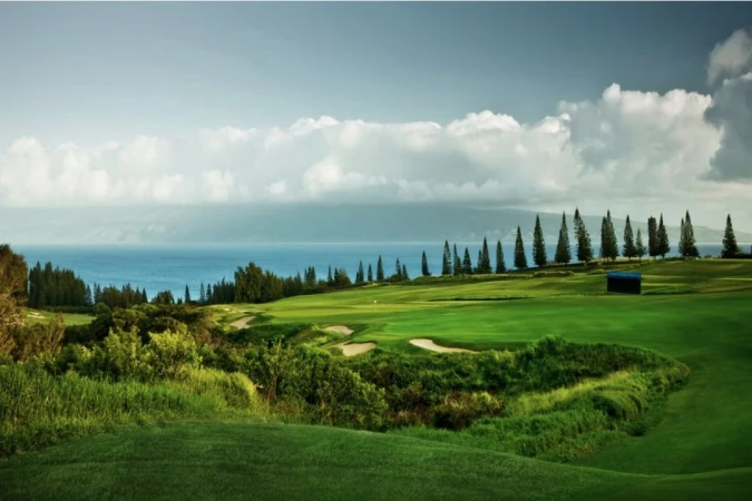 Scenic view of a lush green golf course overlooking the ocean with trees, sand traps, and clouds in the sky.