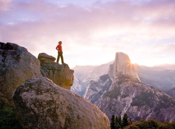 Hiker in red jacket standing on a rocky cliff overlooking a vast mountain valley at sunset by photographer Andy Mahr