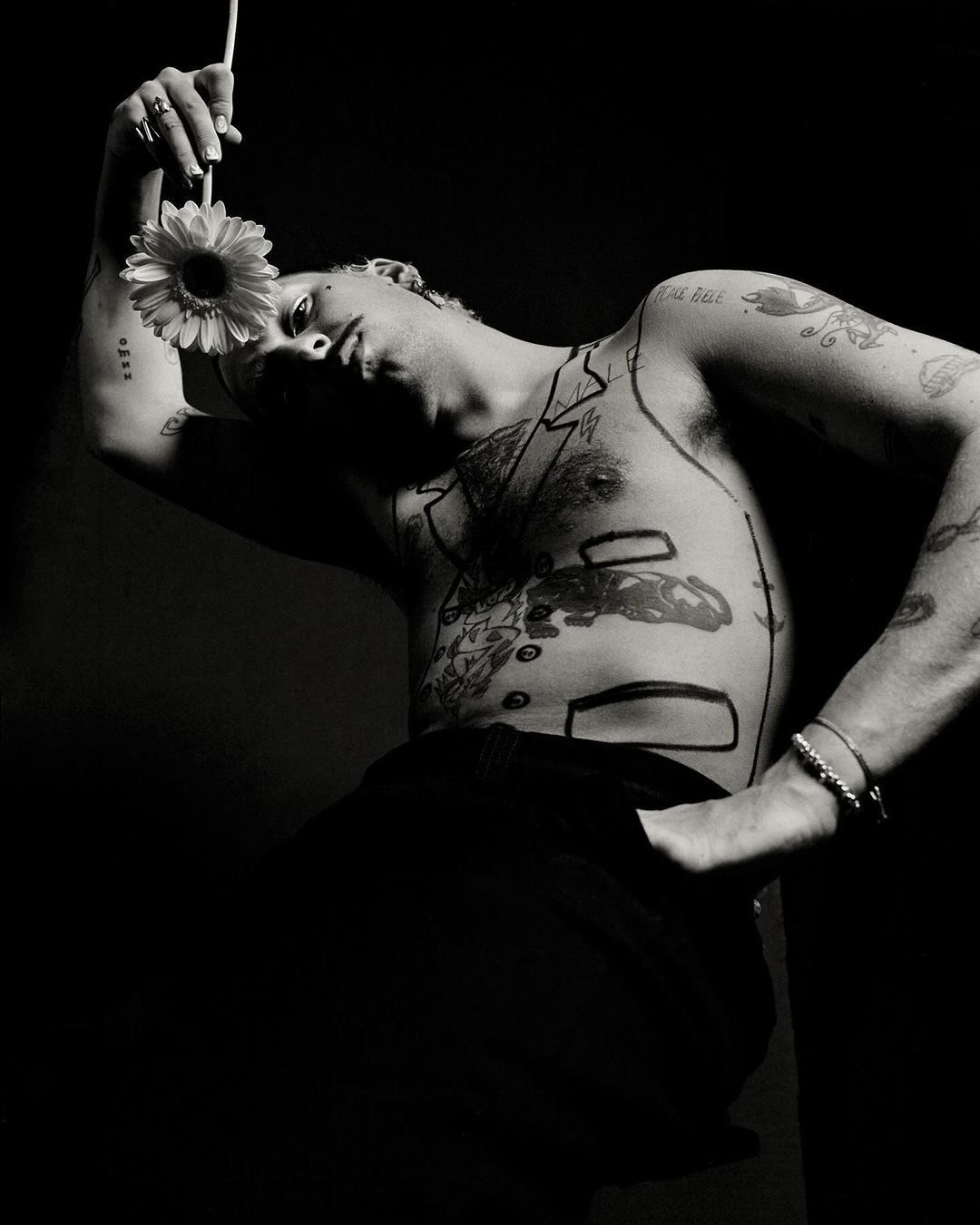 Monochrome portrait of a shirtless adult male with tattoos and drawn-on suit, holding a daisy against a dark background.