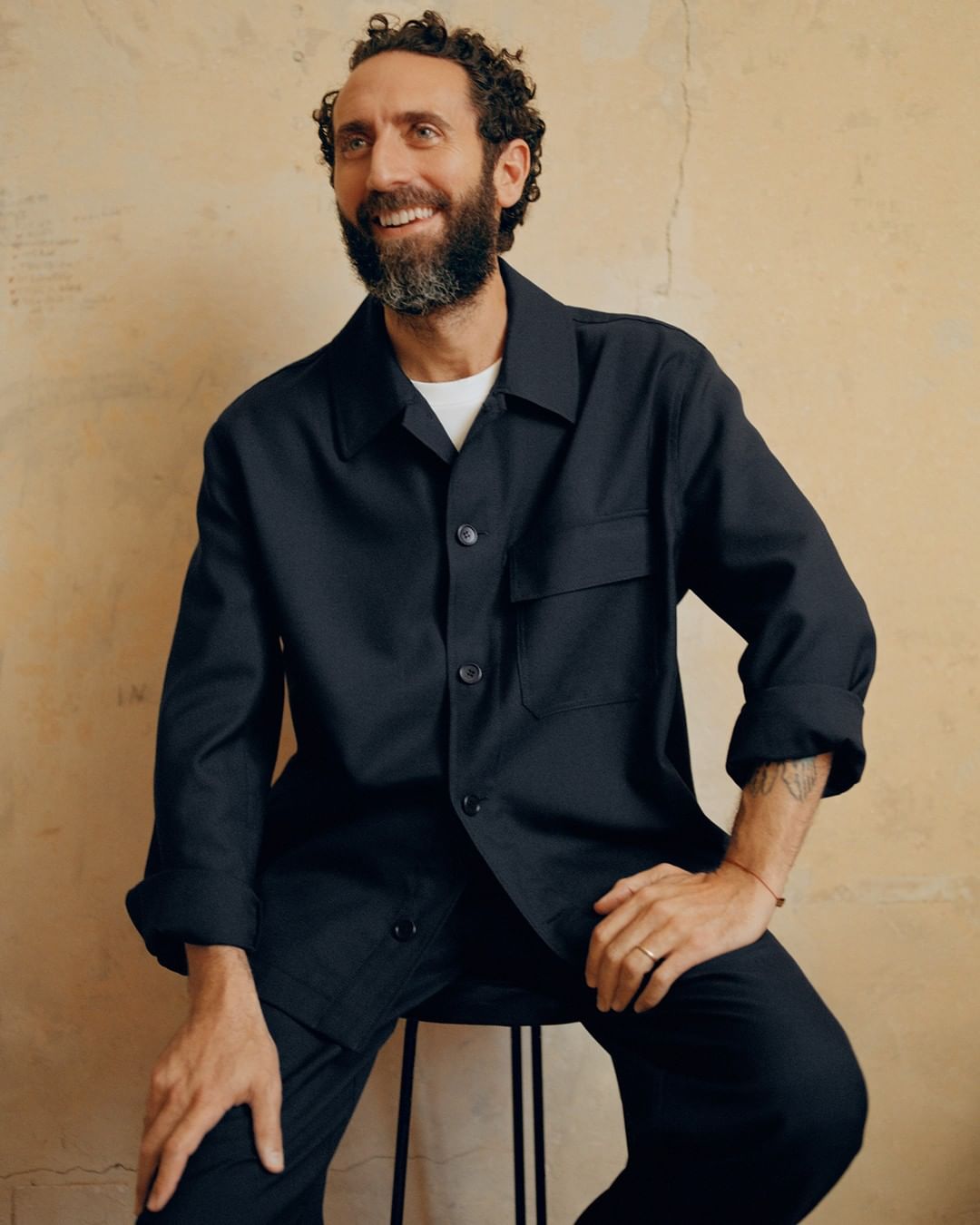 Man with a beard smiles while seated, wearing a dark blue shirt and pants against a neutral backdrop. Natural lighting highlights his relaxed pose.