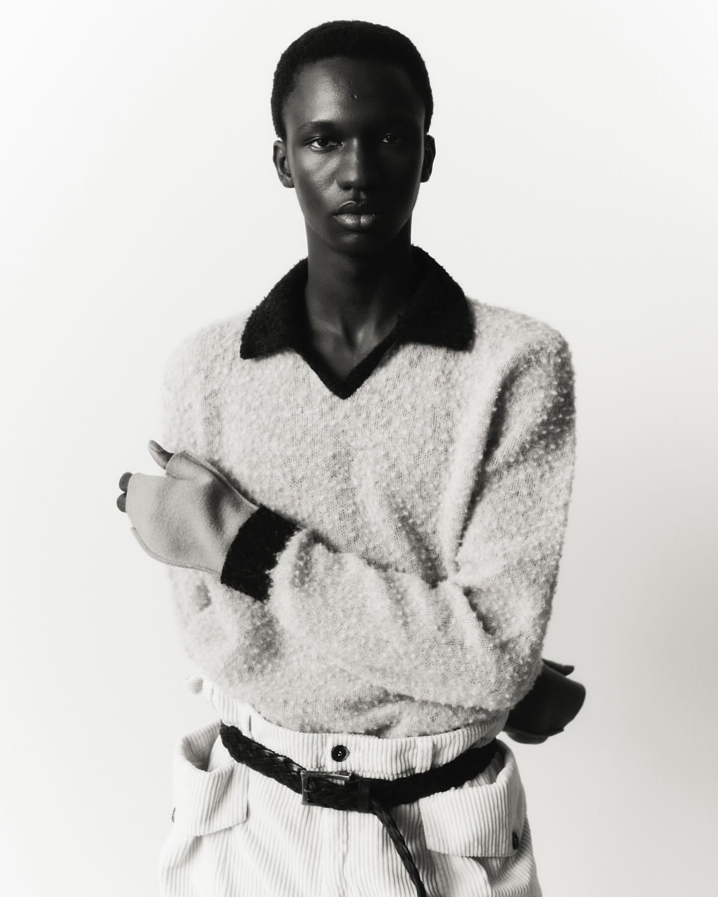 Portrait of a young adult man in a cream-colored sweater, corduroy pants, and black belt, standing against a white backdrop in a photo studio.