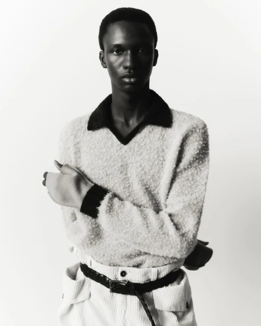 Portrait of a young adult man in a cream-colored sweater, corduroy pants, and black belt, standing against a white backdrop in a photo studio.