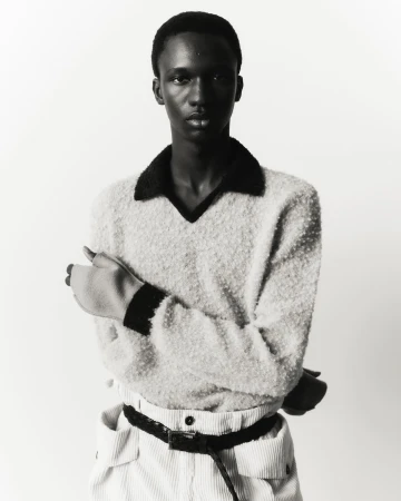 Portrait of a young adult man in a cream-colored sweater, corduroy pants, and black belt, standing against a white backdrop in a photo studio.