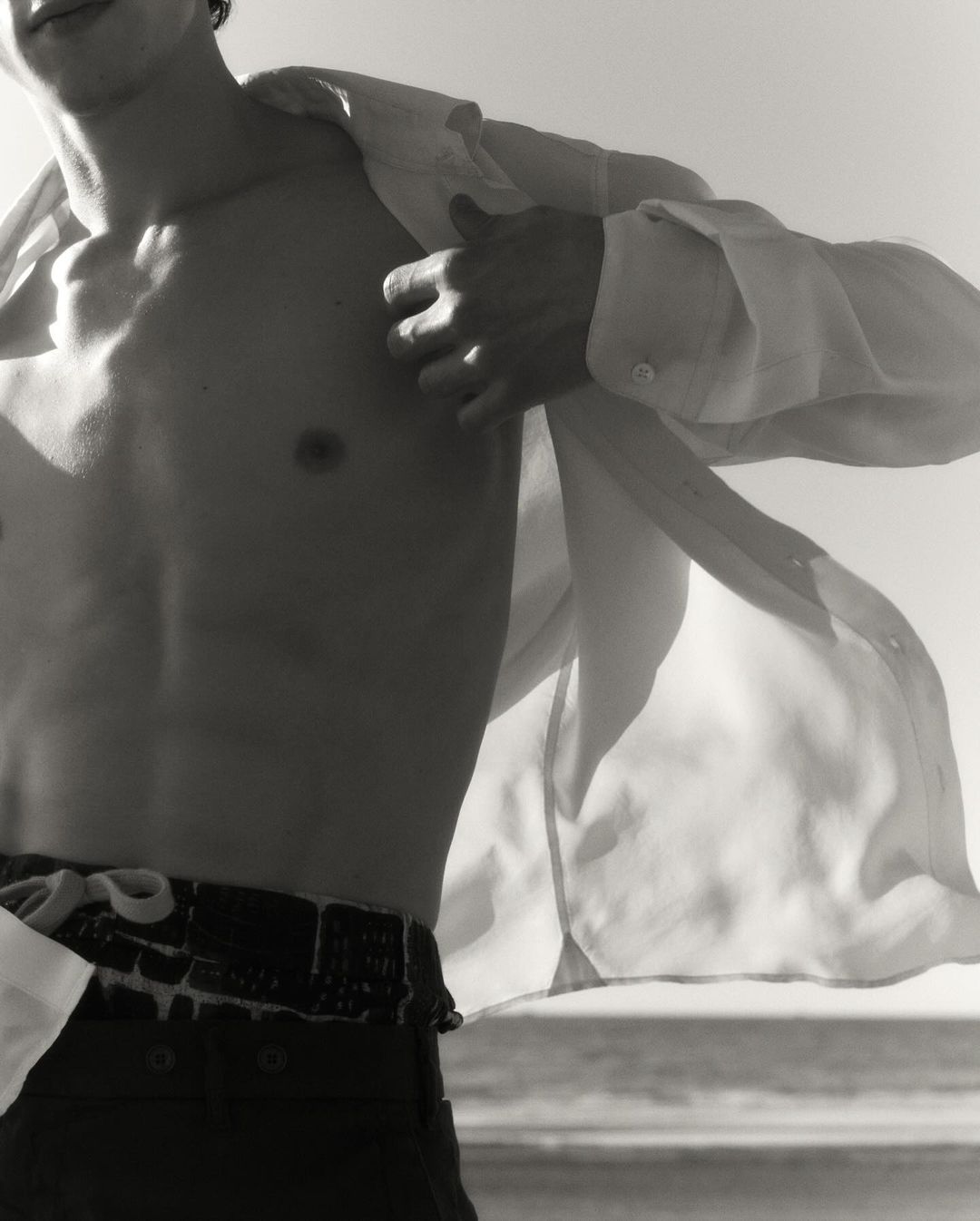 Monochromatic close-up shot of a man with an open shirt revealing his torso against a bright, washed-out background with a glimpse of the sea.