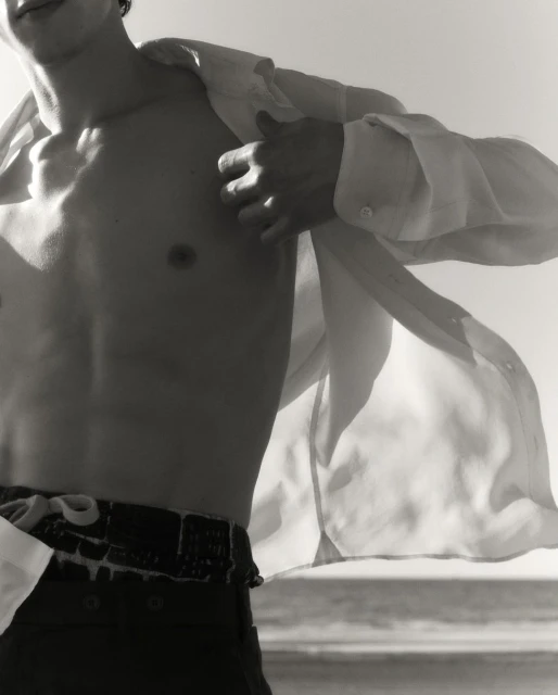 Monochromatic close-up shot of a man with an open shirt revealing his torso against a bright, washed-out background with a glimpse of the sea.