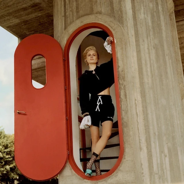 Fashion shot of a blonde woman in a black cropped hoodie and shorts posing in the doorway of a concrete building on stairs.