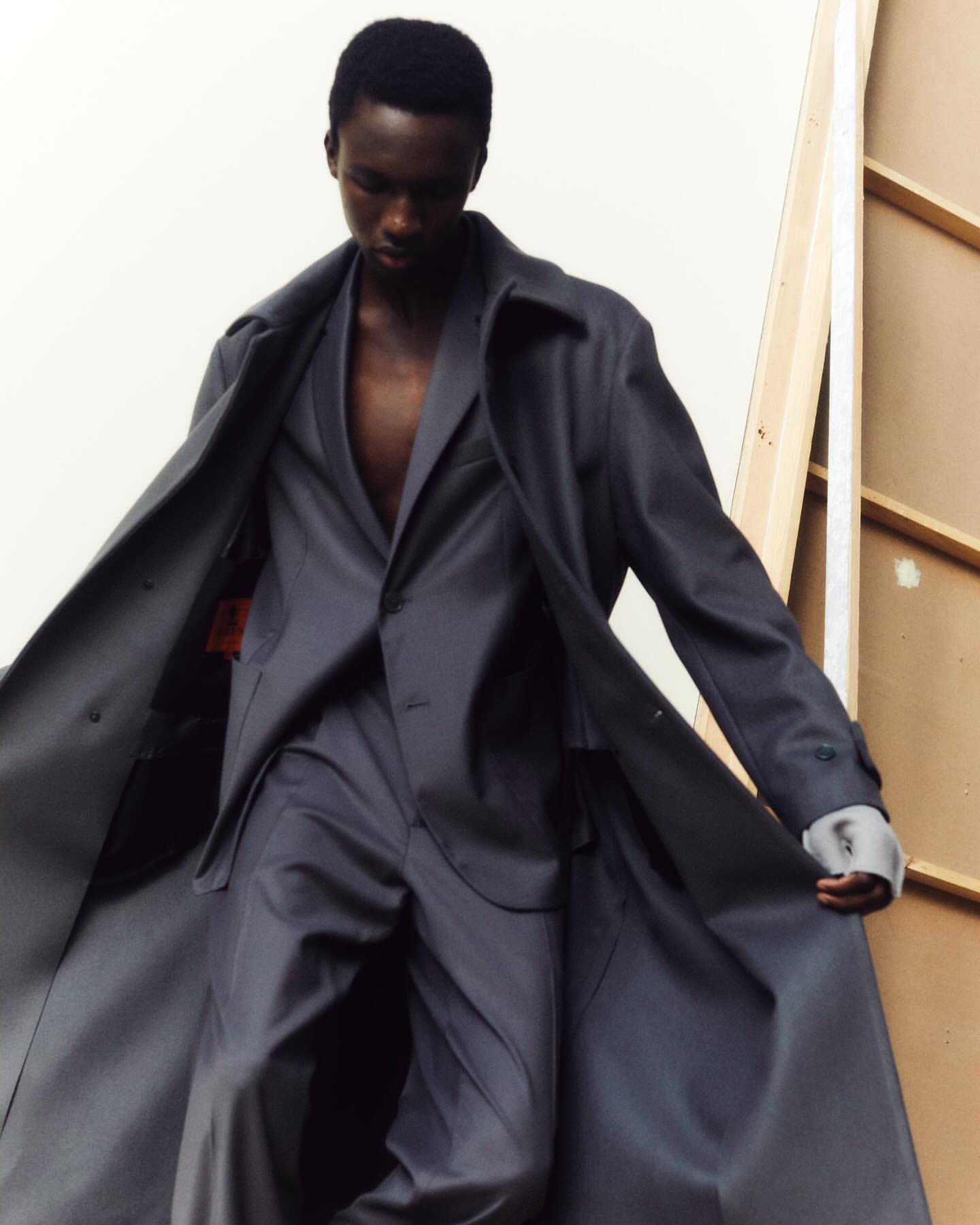 An adult Black male wearing a dark grey suit and coat poses indoors in soft light, looking down, with white wall background.