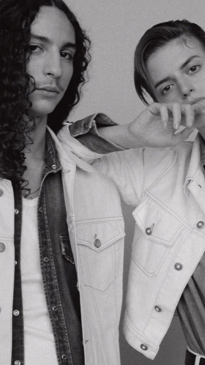 Black and white studio portrait of two adult males posing in denim jackets. One has long curly hair, the other has short slicked-back hair.