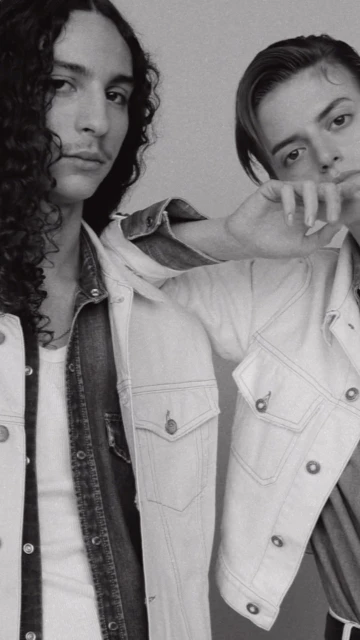 Black and white studio portrait of two adult males posing in denim jackets. One has long curly hair, the other has short slicked-back hair.