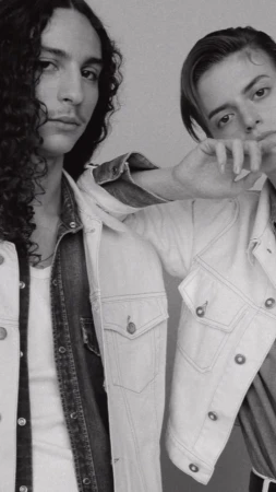 Black and white studio portrait of two adult males posing in denim jackets. One has long curly hair, the other has short slicked-back hair.
