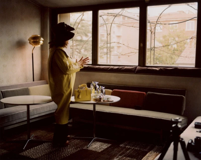 Woman in yellow coat and large hat stands near a window in a room with couches, tables, and lamps, creating a vintage aesthetic.