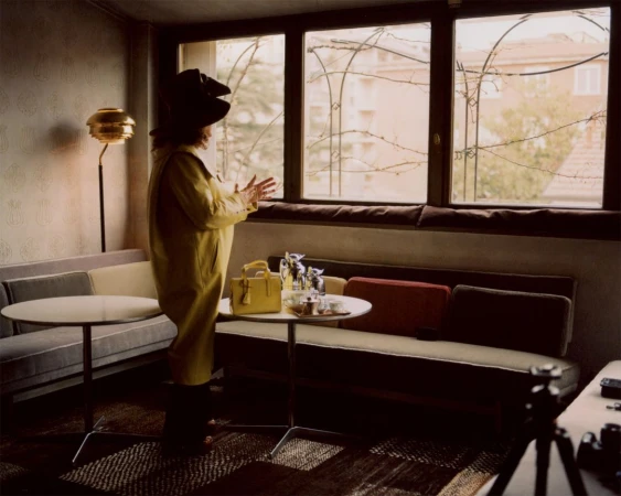 Woman in yellow coat and large hat stands near a window in a room with couches, tables, and lamps, creating a vintage aesthetic.