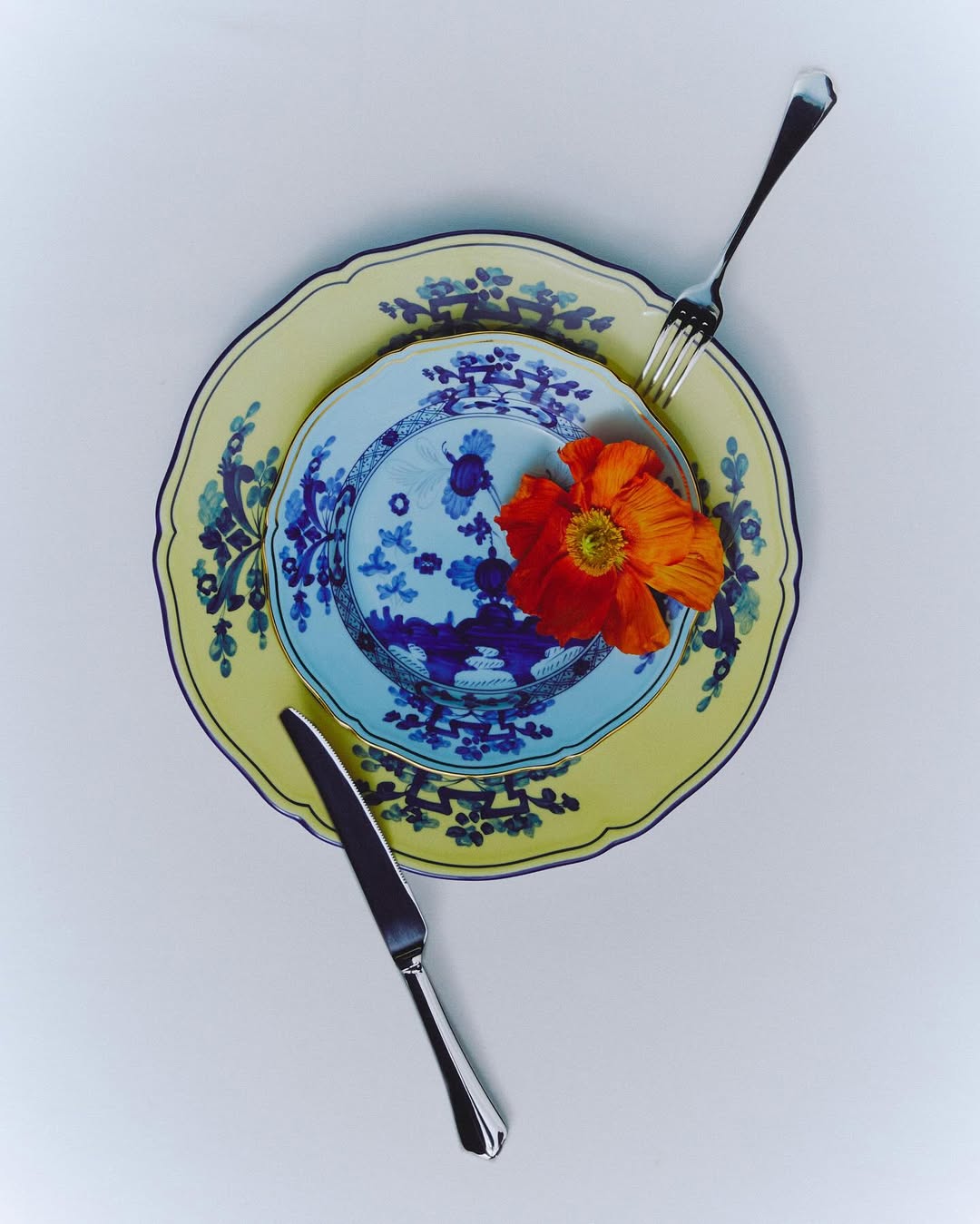 Overhead shot of layered yellow and blue patterned plates with a knife, fork, and an orange poppy flower against a white backdrop.