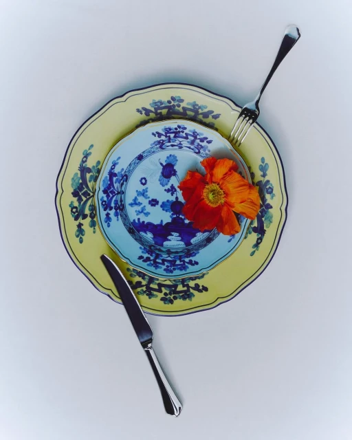 Overhead shot of layered yellow and blue patterned plates with a knife, fork, and an orange poppy flower against a white backdrop.