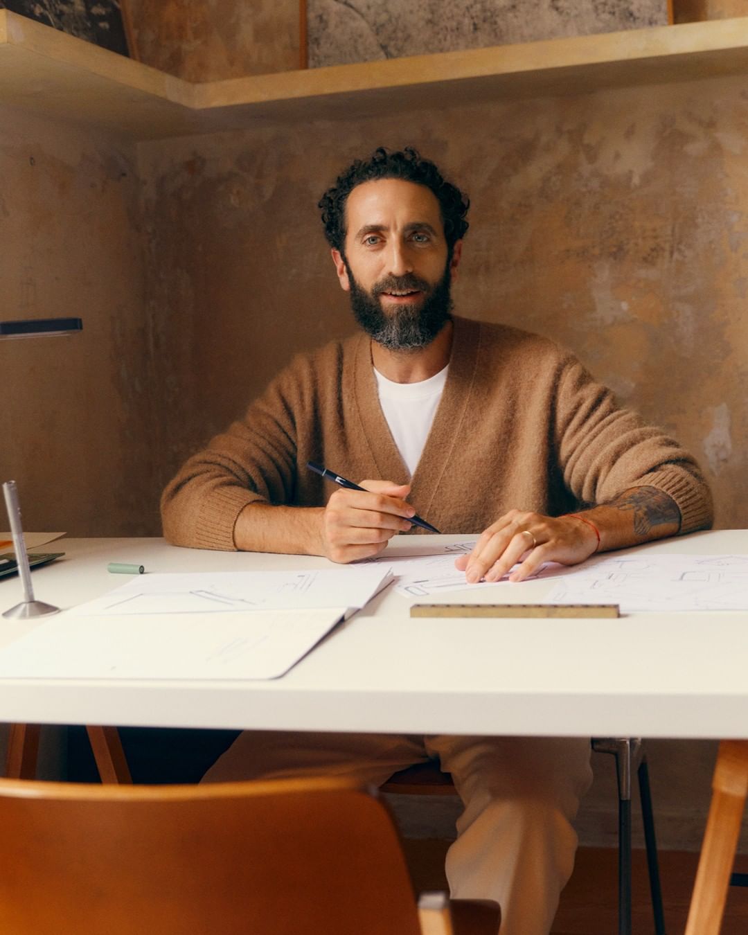Bearded man in cardigan at desk working on architectural designs in room with neutral tones and modern lamp.