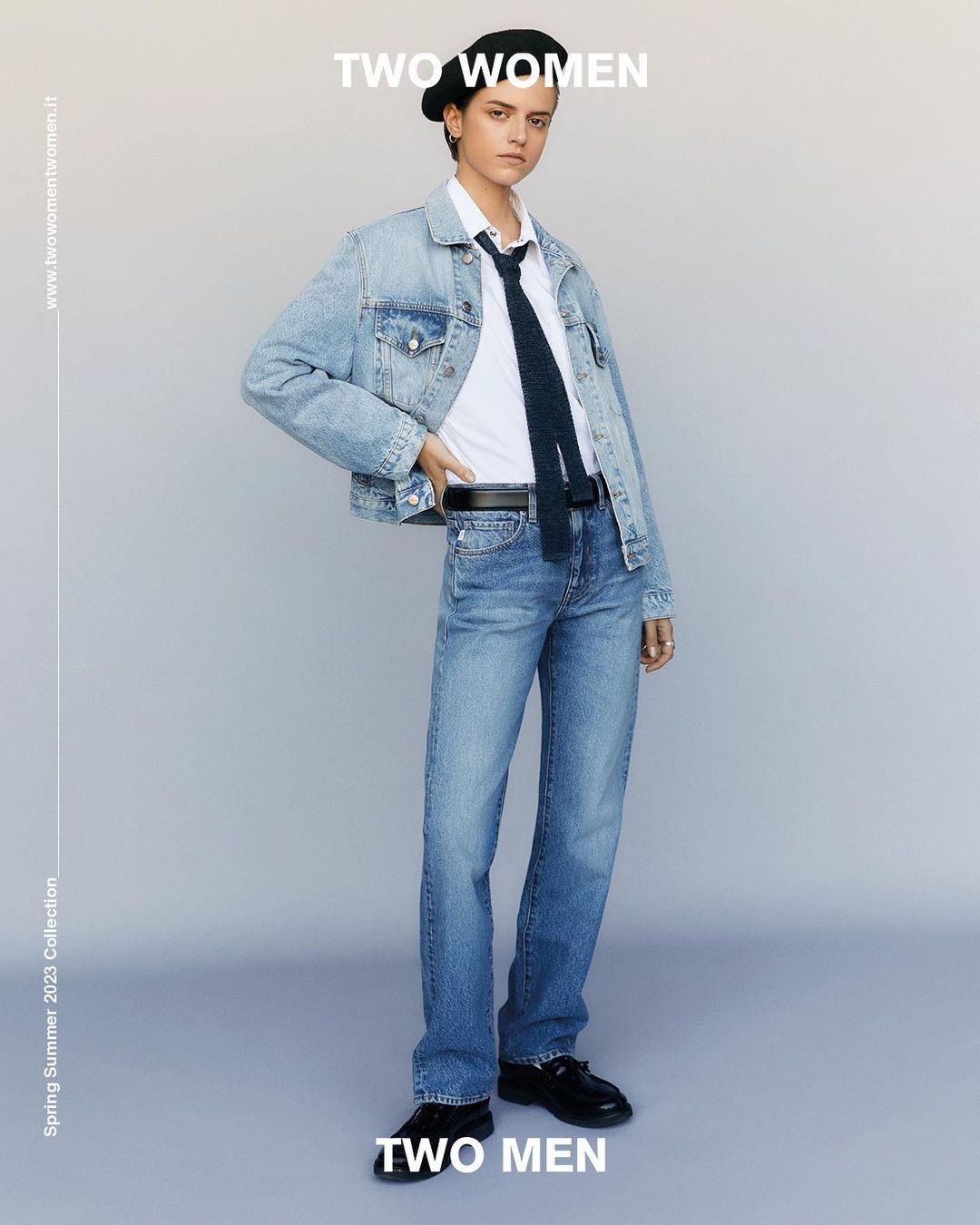 Fashion shot of an androgynous person in a denim jacket, white shirt, jeans, beret, and tie standing in a photo studio.
