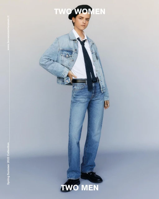 Fashion shot of an androgynous person in a denim jacket, white shirt, jeans, beret, and tie standing in a photo studio.