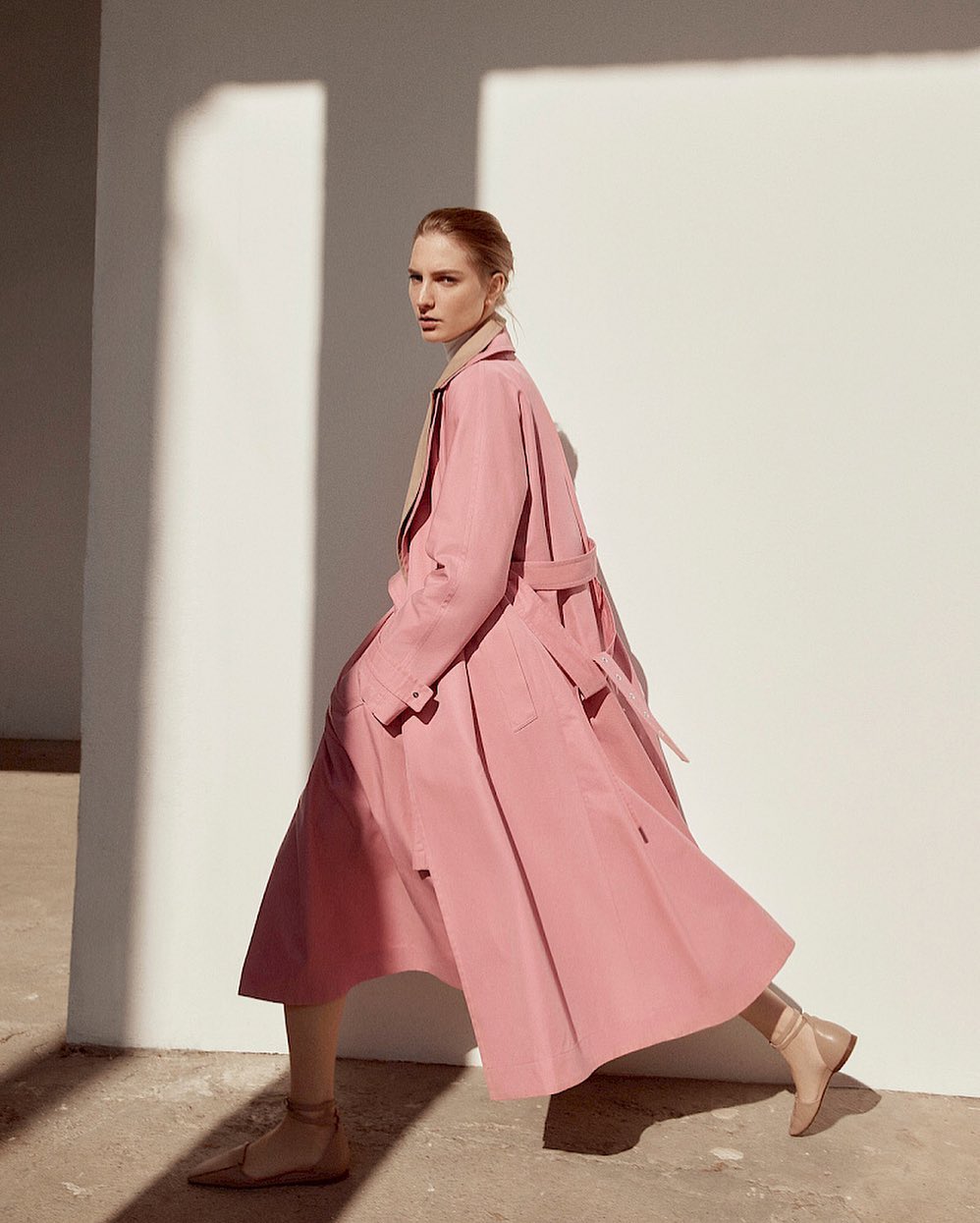 Elegant woman in pink trench coat and beige ankle boots walks against a white wall with hard light casting geometric shadows.