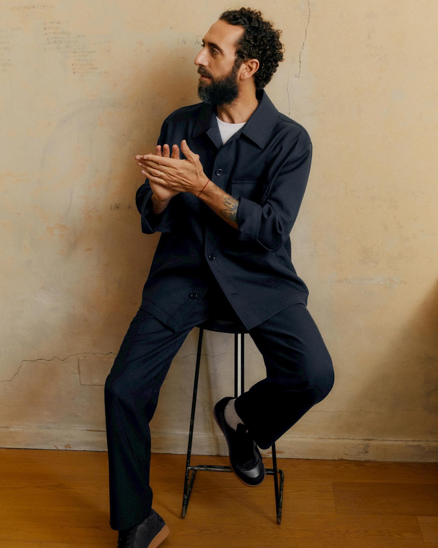 Bearded adult man wearing a navy buttoned shirt and matching pants sitting on a stool, looking off to the left in an indoor setting.